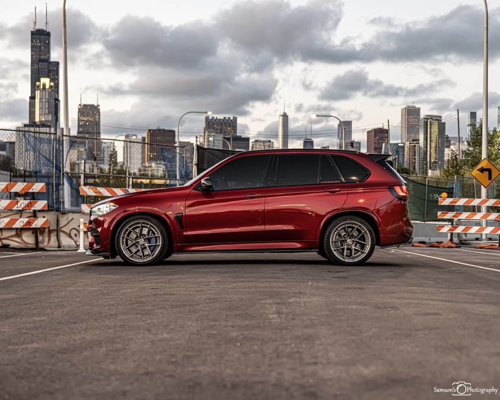 Red luxury SUV parked on a roof with a city skyline in the background under a cloudy sky.